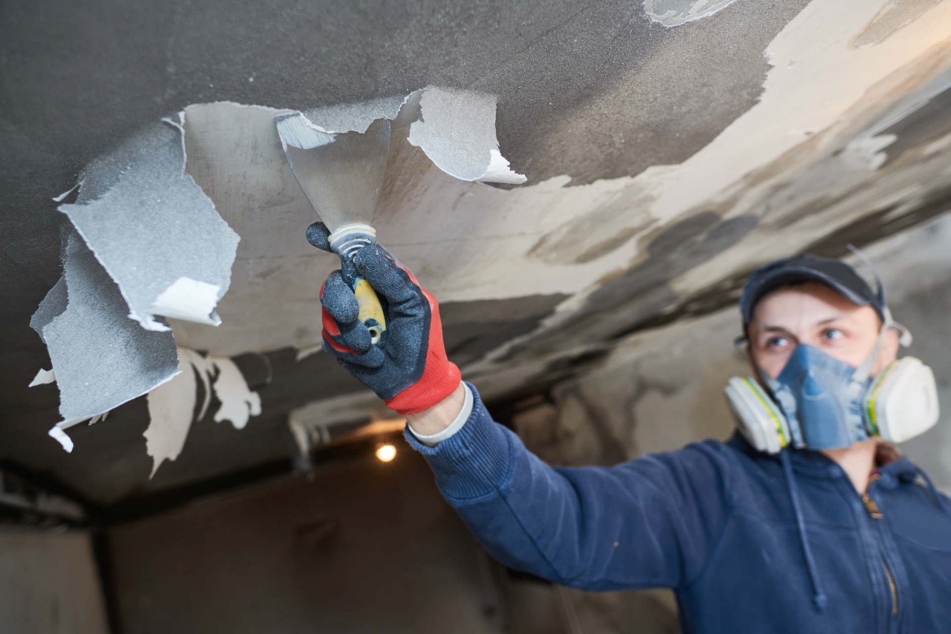 Worker removes damaged paint layer from wall and ceiling indoors.